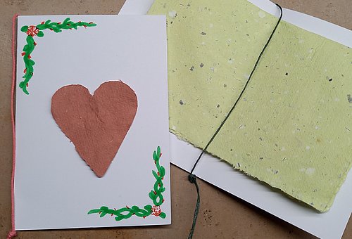 Zwei Karten aud weißem Karton wurden mit selbst geschöpftem, rauem Papier verziert. Eine Karte zeigt außen ein rotes Herz aus Papier und gezeichnete, grüne Ranken. In die zweite Karte wurde innen ein hellgrünes, geflecktes Stück selbst geschöpftes Papier gelegt und mit einem Faden festgebunden.